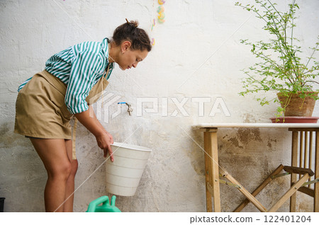 Woman Filling a Bucket from an Outdoor Tap Near a Gardening Area 122401204