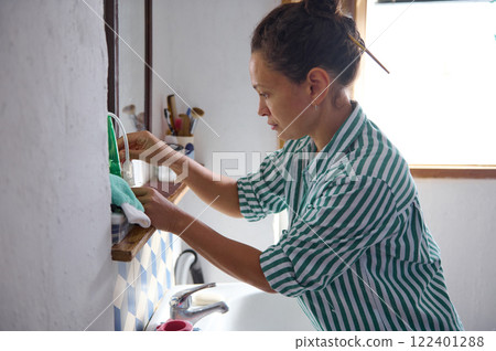 Woman cleaning a bathroom mirror while organizing a sink area 122401288