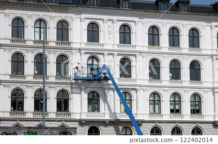 Worker paints the facade of the building at heigt in lifting cradle. Construction workers at height working on crane in lifting bucket. Worker paints the facade of the building at heigt in lifting cradle. Construction workers at height working on crane in lifting bucket. 122402014