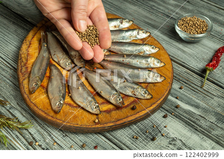 A hand delicately sprinkles aromatic spices or coriander on a round wooden board adorned with freshly washed fish. The rustic kitchen setting evokes culinary charm, attracting the taste buds 122402999