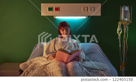 Positive and relaxed young woman in hospital gown lying in bed and reading book in the evening after her procedures in well-equipped hospital ward in clinic Positive and relaxed young woman in hospital gown lying in bed and reading book in the evening after her procedures in well-equipped hospital ward in clinic 122403150