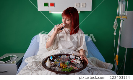 Young red-haired woman sitting in bed in hospital ward with tray filled with various meds and using nasal spray against green wall in modern clinic Young red-haired woman sitting in bed in hospital ward with tray filled with various meds and using nasal spray against green wall in modern clinic 122403154