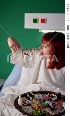 Young red-haired woman sitting in bed with tray filled with many different meds and taking pills with large spoon. Recovery process in modern clinic 122403155