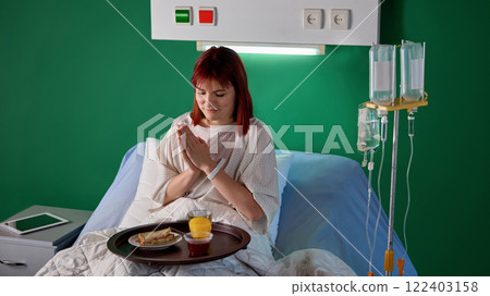 Positive young red-haired woman in medical gown sitting in bed in hospital ward with tray of delicious food and preparing to have dinner in modern clinic 122403158