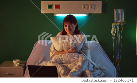Young emotionally woman in hospital gown sitting in bed in hospital ward in evening, hugging pillow and crying while watching dramatic and sad movie on her laptop 122403171