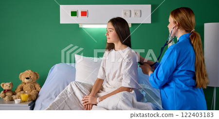 Teen girl sitting on bed in hospital ward while female nurse examining her check breath sound with stethoscope in modern clinic Teen girl sitting on bed in hospital ward while female nurse examining her check breath sound with stethoscope in modern clinic 122403183