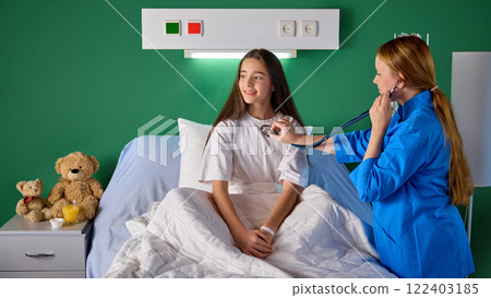 Teen girl sitting on bed in hospital ward while female nurse examining her check breath sound and heartbeat with stethoscope in modern clinic 122403185