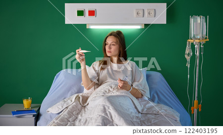 Young woman in white gown sitting in bed in comfortable and spacious hospital ward and measuring temperature against green wall 122403195