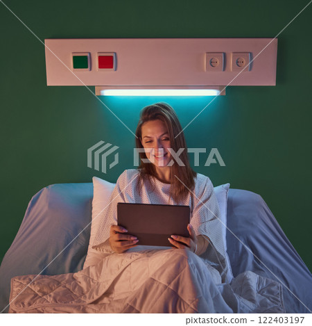 Young woman in comfortable gown sitting in bed in her hospital ward in evening and using tablet. Recovery process, relaxation time 122403197