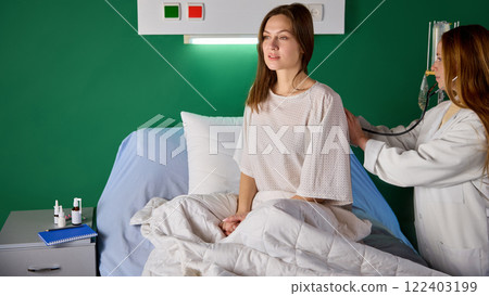 Young woman sitting in bed in hospital gown in hospital ward while female nurse checking her breath and heartbeat with stethoscope 122403199