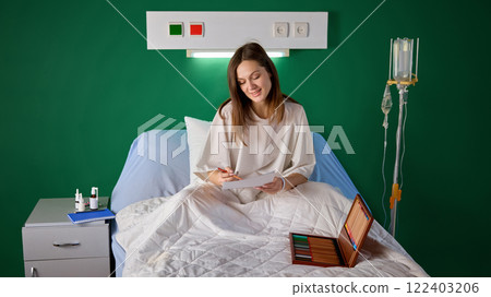 Young woman in hospital gown sitting in bed in her hospital ward and drawing on paper with colorful crayons placed in wooden box 122403206