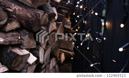 Neatly stacked firewood for the winter heating season arranged along a black fence decorated with glowing Christmas lights creating a cozy and festive outdoor scene perfect for the holiday atmosphere 122403521