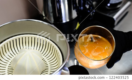 A glass of freshly squeezed citrus juice from ripe oranges served with ice in a clear glass placed next to a juicer held by a barista in a modern cafe setting emphasizing freshness and health 122403525