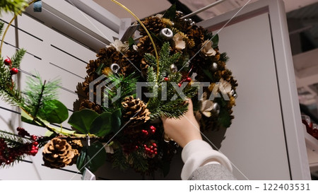 A woman carefully selects holiday home decor from a shop, choosing festive wreaths with pinecones and artificial pine branches, creating a cozy Christmas atmosphere. 122403531