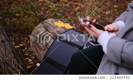 A woman in a gray coat charges her phone using a portable solar panel in a serene forest setting. The scene highlights sustainable technology, off-grid living, and eco-friendly solutions in nature. 122403541