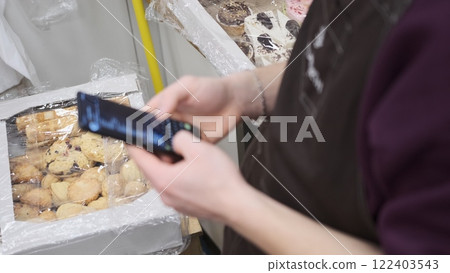 Out of focus, a vendor wearing a brown apron uses a smartphone to order a new stock of sweets and cookies for sale, creating a casual scene of business management in a cozy and inviting shop setting. Out of focus, a vendor wearing a brown apron uses a smartphone to order a new stock of sweets and cookies for sale, creating a casual scene of business management in a cozy and inviting shop setting. 122403543