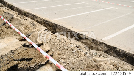A freshly dug trench along a parking lot, marked with red and white striped safety tape. The scene highlights active construction work and safety measures, showcasing urban development in progress. 122403569