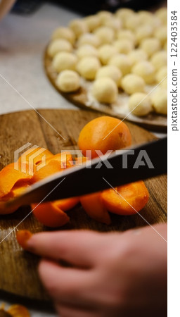 Hands of a woman delicately slice ripe apricots and bananas on a rustic wooden plate, featuring lazy dumplings in the background. Ideal for food enthusiasts seeking culinary inspiration vertical 122403584