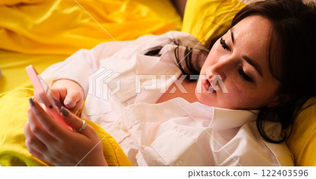 A young Caucasian woman in a white T-shirt uses a smartphone while lying on a bed with yellow bedsheets. Suitable for lifestyle, technology, and relaxation-themed videos. 122403596
