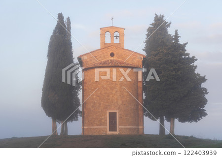Small, old stone chapel stands enveloped in morning fog, surrounded by tall cypress trees in Tuscany 122403939