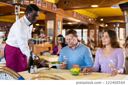 Polite african american waiter serving dishes to positive couple in restaurant 122408584