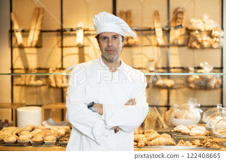 Confident adult man, bakery owner in white chef jacket and cap looking at the camera during working day in bakehouse. Cooking, culinary and bakery concept 122408665
