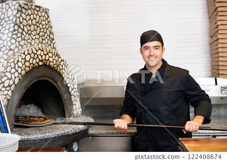 Latino chef in black uniform holding shovel with pizza in kitchen restaurant Latino chef in black uniform holding shovel with pizza in kitchen restaurant 122408674