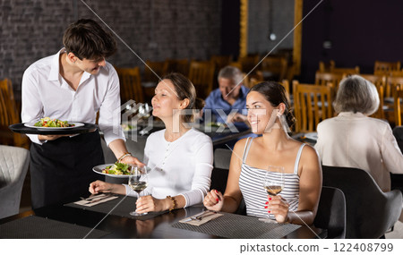 Young male waiter serving dish to two women in restaurant Young male waiter serving dish to two women in restaurant 122408799