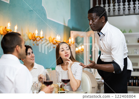 Positive couple talking with polite african american waiter while dining in cozy restaurant Positive couple talking with polite african american waiter while dining in cozy restaurant 122408804