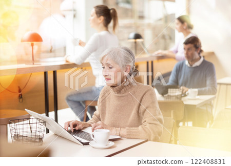 Elderly woman sitting at table, using laptop and enjoing cup of tea in coffee house 122408831