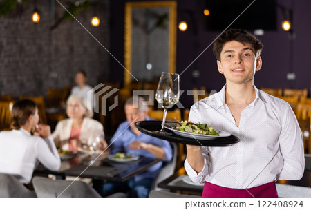 Young waiter guy is standing with order tray in spacious guest room in restaurant. 122408924