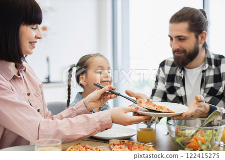Caucasian family having breakfast with pizza and salad. Young parents with daughter sharing meal. Bright kitchen setting, creating joyful and warm family atmosphere. Caucasian family having breakfast with pizza and salad. Young parents with daughter sharing meal. Bright kitchen setting, creating joyful and warm family atmosphere. 122415275