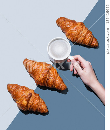 Hand holding cup of cappuccino or latte coffee on blue background with fresh croissants and shadow. Hand holding cup of cappuccino or latte coffee on blue background with fresh croissants and shadow. 122419653