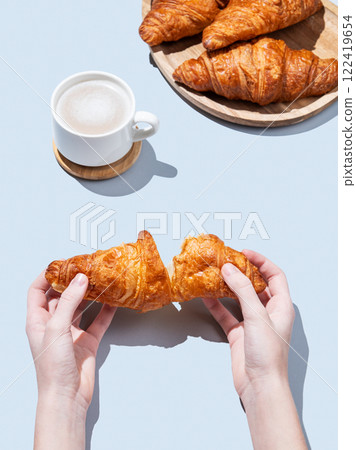 Hands breaking a fresh croissant over a blue background with a cup of cappuccino or latte coffee Hands breaking a fresh croissant over a blue background with a cup of cappuccino or latte coffee 122419654