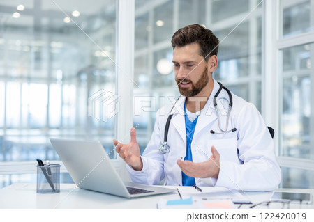 Doctor inside clinic office using laptop for video call, man in white medical coat talking remotely, consulting patients while sitting at desk inside office. 122420019