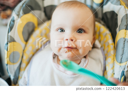 Baby sitting in a high chair with food on their face and a spoon being offered with a Messy Face While Eating. Early feeding and childcare concept 122420613