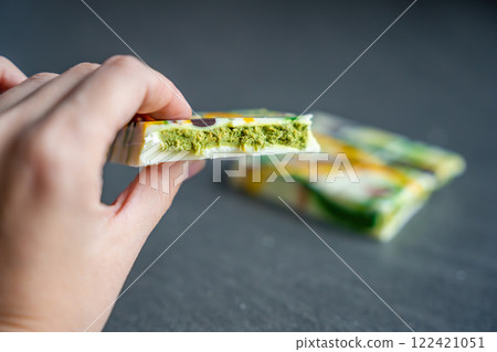 Piece of trend Dubai white chocolate with pistachio paste and kataifi dough in woman hand. Confectionery handmade sweets at home in the kitchen. Piece of trend Dubai white chocolate with pistachio paste and kataifi dough in woman hand. Confectionery handmade sweets at home in the kitchen. 122421051
