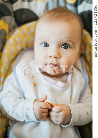 Cute Baby with Messy Face Eating Solid Food During Mealtime. Baby sitting in a high chair with food on their face, holding a piece of food. Early self-feeding and baby nutrition concept Cute Baby with Messy Face Eating Solid Food During Mealtime. Baby sitting in a high chair with food on their face, holding a piece of food. Early self-feeding and baby nutrition concept 122421650