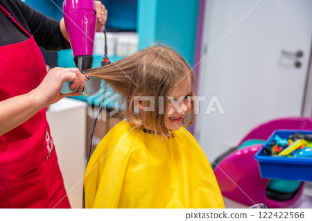 Hairdresser making a hair style to cute little girl in real child salon with colorful interior and toys 122422566