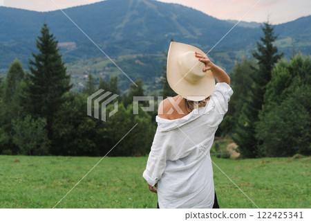 Young woman in hat standing on green meadow in mountains, rear view 122425341