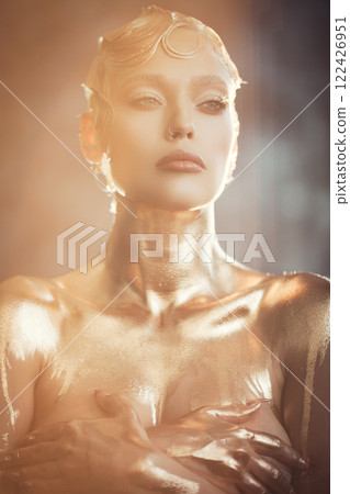 Beauty portrait of young attractive fashion model with short wet blond hair. Golden paint on body. Blue eyes, professional makeup. Warm colors. Natural sunlight and shadows on background indoors 122426951