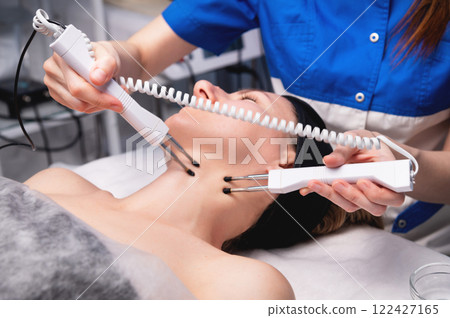 Happy woman gets ultrasonic facial massage with ultrasonic facial machine. Woman undergoing electric facelift procedure in luxury beauty salon 122427165
