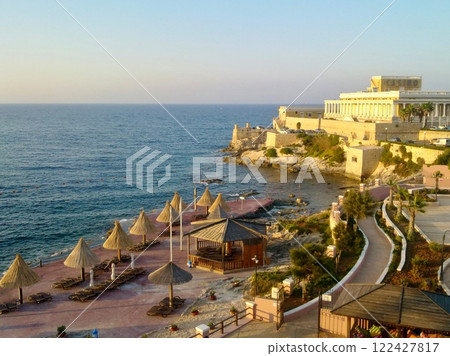 A scenic coastal view featuring a Mediterranean seaside with straw umbrellas, sun loungers, and a historic casino building overlooking the clear blue sea. A scenic coastal view featuring a Mediterranean seaside with straw umbrellas, sun loungers, and a historic casino building overlooking the clear blue sea. 122427817