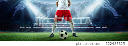 Dynamic soccer moment. Soccer player in red shorts standing with ball at 3D model of stadium under bright spotlights. 122427825