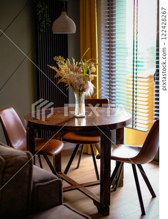 A table with four chairs and a vase of flowers in the center. The table is surrounded by a couch and a window A table with four chairs and a vase of flowers in the center. The table is surrounded by a couch and a window 122428267
