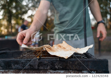 A man checks grilled meat and flatbread on a barbecue, cooking outdoors amidst trees in the evening A man checks grilled meat and flatbread on a barbecue, cooking outdoors amidst trees in the evening 122428348