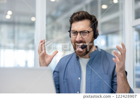 Angry angry man shouting while sitting with headset and laptop inside office, video call of office worker. Angry angry man shouting while sitting with headset and laptop inside office, video call of office worker. 122428907