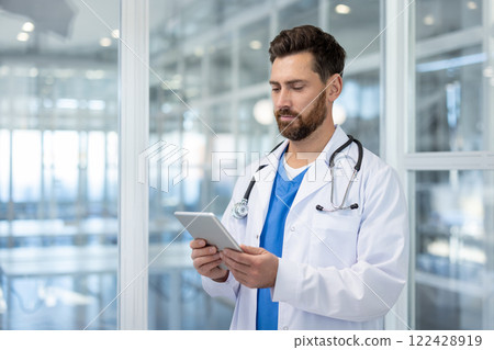 Serious experienced doctor standing inside the clinic with a tablet computer in his hands. Mature man in a white medical coat and with a stethoscope using an application. Serious experienced doctor standing inside the clinic with a tablet computer in his hands. Mature man in a white medical coat and with a stethoscope using an application. 122428919