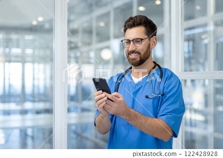 Male doctor wearing blue scrubs and stethoscope uses smartphone in modern healthcare setting, conveying professionalism and technology. Represents healthcare, communication, and digital innovation. 122428928