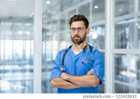 Confident male doctor in blue scrubs with stethoscope, standing in modern medical facility. Medical professional with glasses portrays expertise and trust in healthcare setting. 122428933